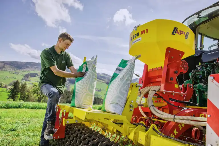 Landwirt mit Saatgut-Säcken vor der Nachsaat, Sämaschine und Traktor, im Vordergrund, Hügel und blauer Himmel im Hintergrund, Totale