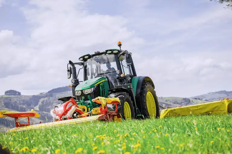 Landwirt im John Deere-Traktor bei der Mahd, Hügel und grauer Himmel im Hintergrund, Totale